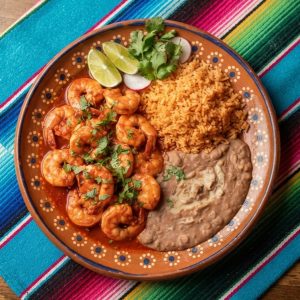 Shrimp in a spicy red chile sauce with Mexican rice and refried beans, on a terracotta clay plate.