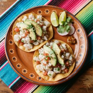 Two flat crisp tostadas topped with lime-cured fish ceviche and sliced avocado, on a terracotta clay plate.