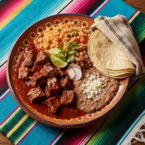 Beef simmered in red chile sauce with Mexican rice, refried beans and warm tortillas, on a terracotta clay plate.