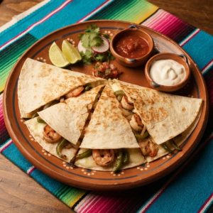 A quesadilla cut into wedges showing shrimp, melted cheese, peppers and onions, on a terracotta clay plate.