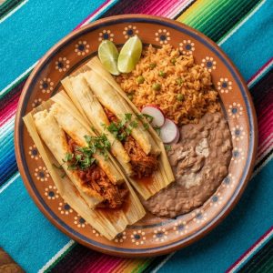 Two tamales with their corn husks opened, showing masa and pork in red chile sauce, with rice and beans, on a terracotta clay plate.